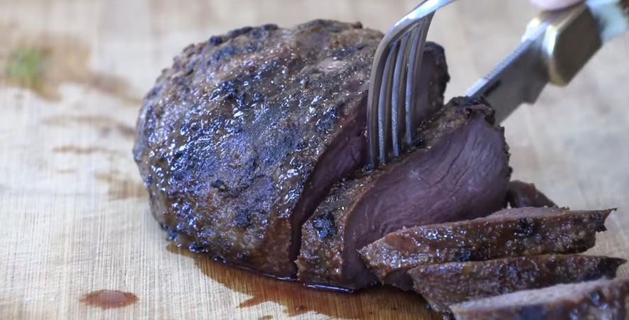 Piece of meat being cut on a cutting board with a knife. It looks like steak. 