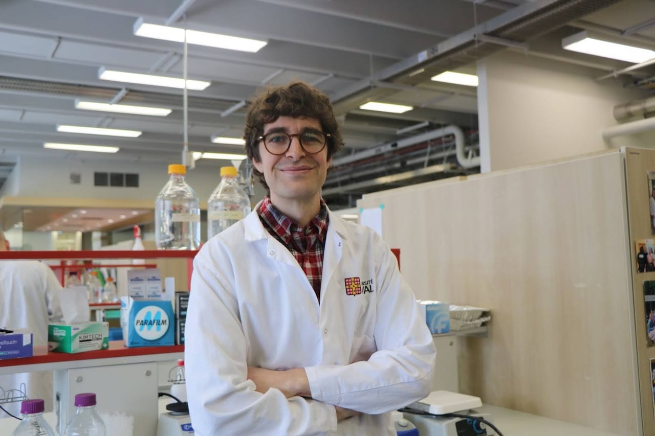 A man in a white lab coat smiling with his arms crossed in a lab. 