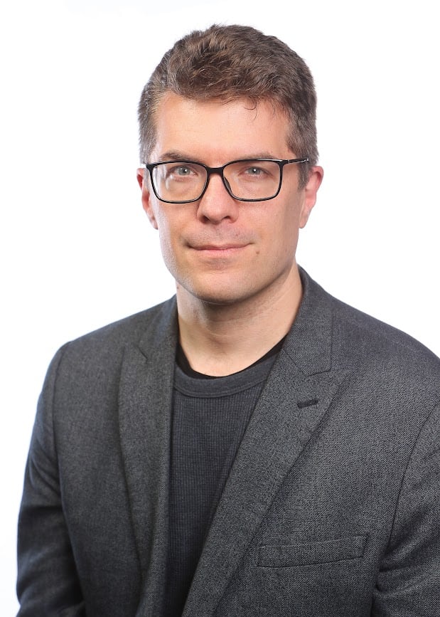 A portrait of a man wearing glasses dressed in dark-grey suits.