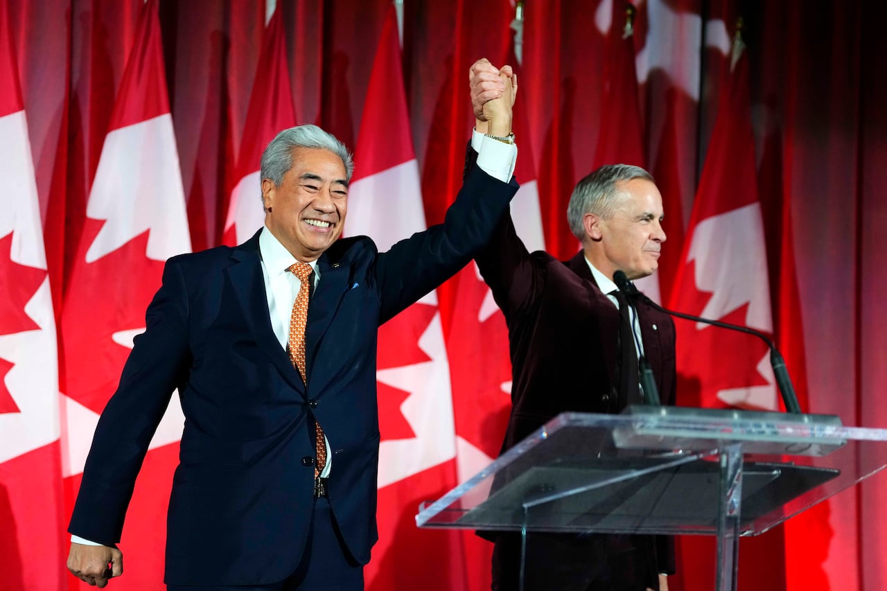 Two men in suits raise their hands together on a stage lined with Canadian flags.