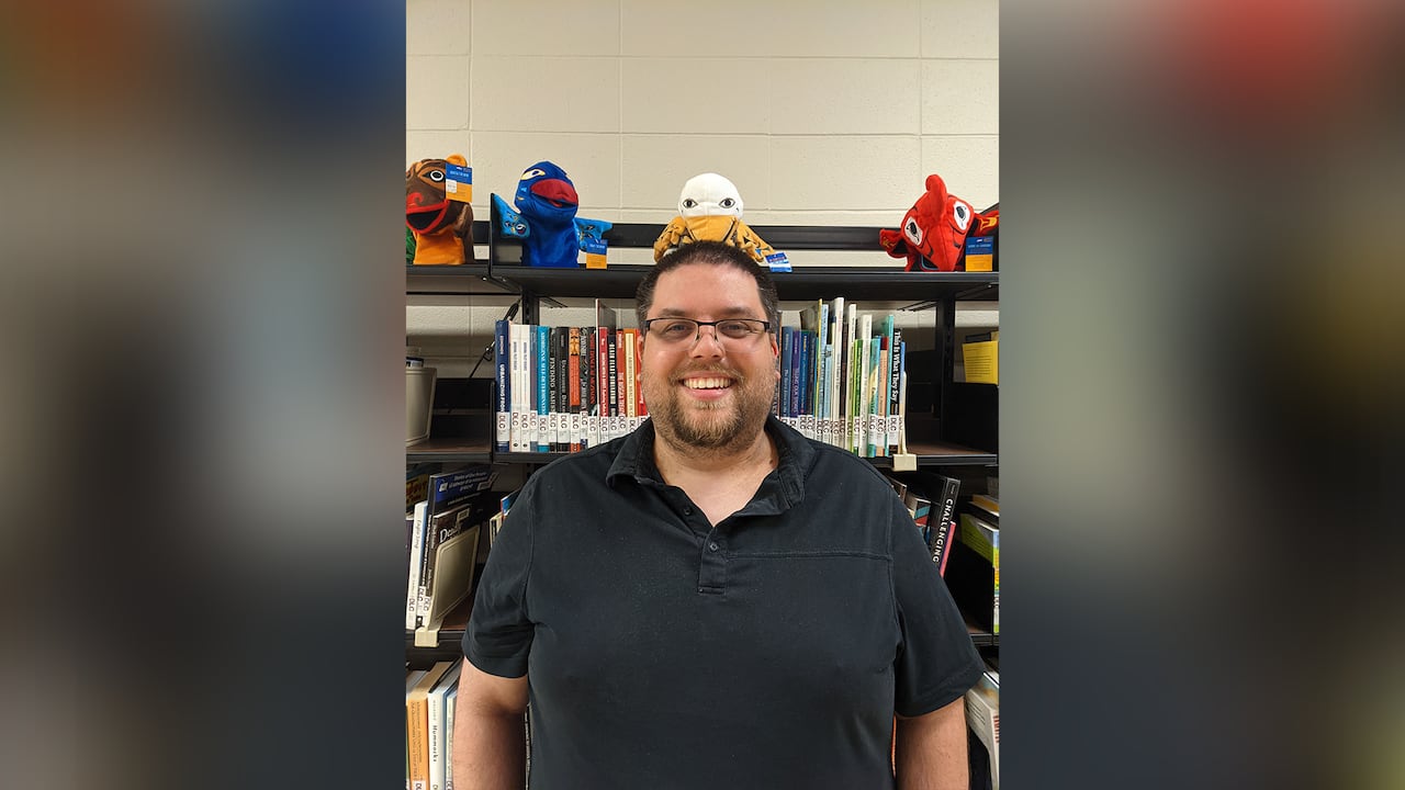 A smiling man in a black golf tee stands before a bookcase full of books, a stuffed doll perched on the shelf above his head.