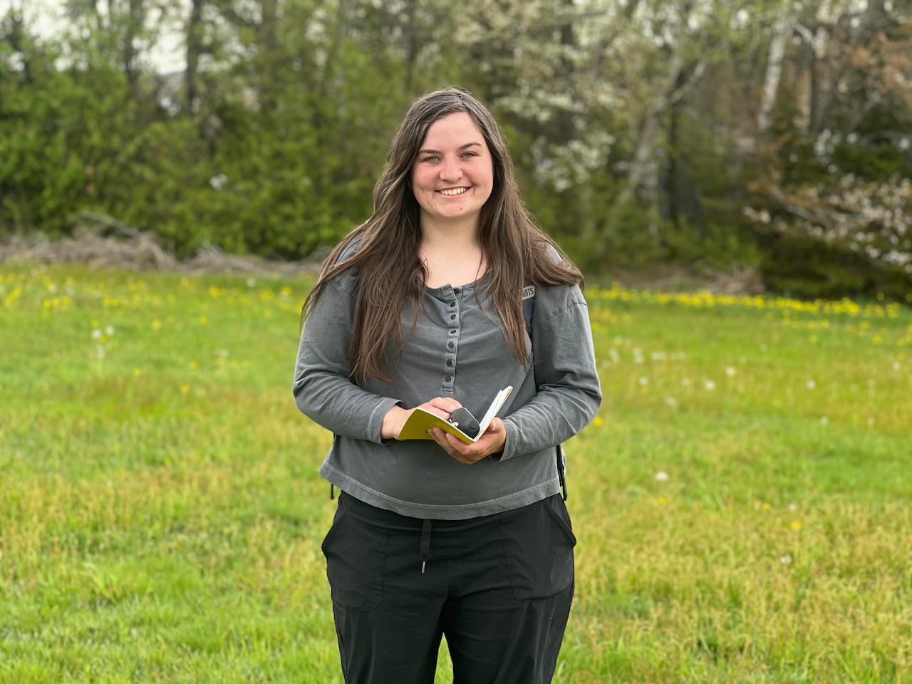 A woman holding a notebook stands on a treed lot 