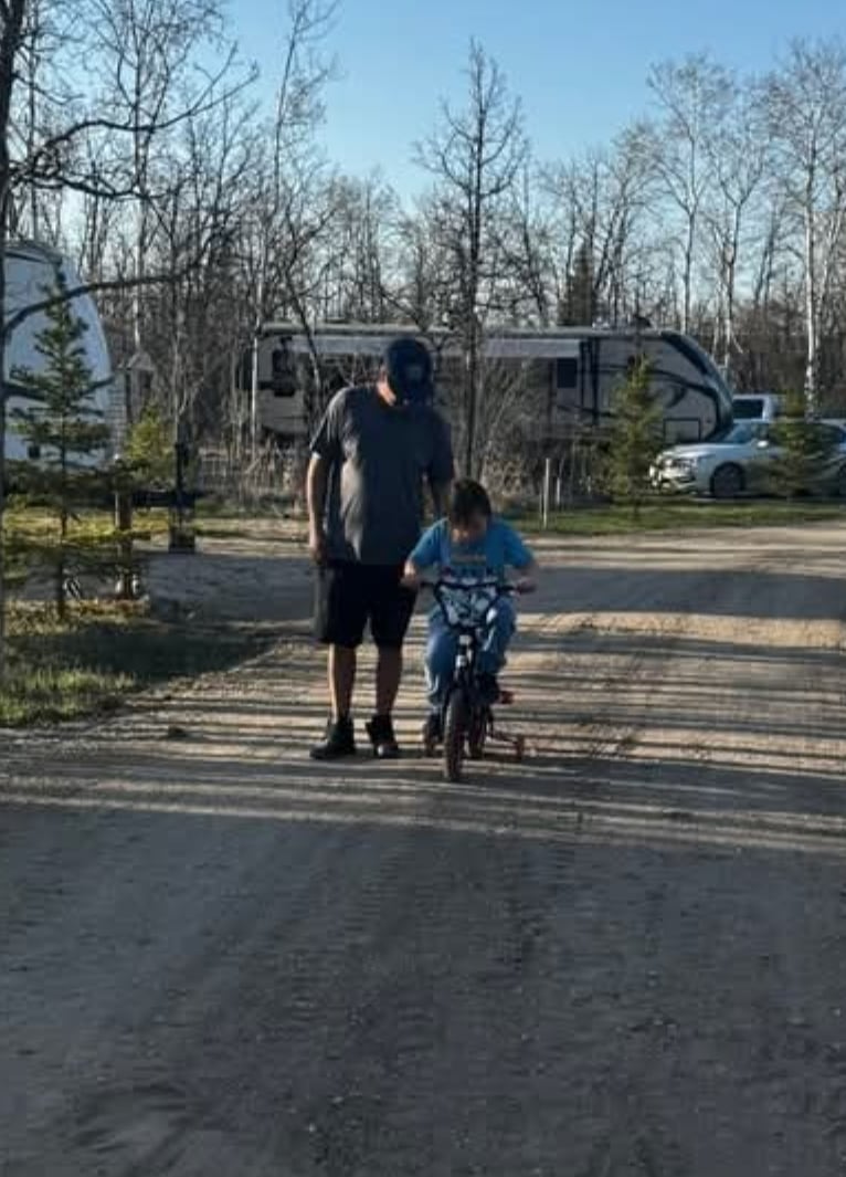 A man walks alongside a boy riding a bike through a campground.
