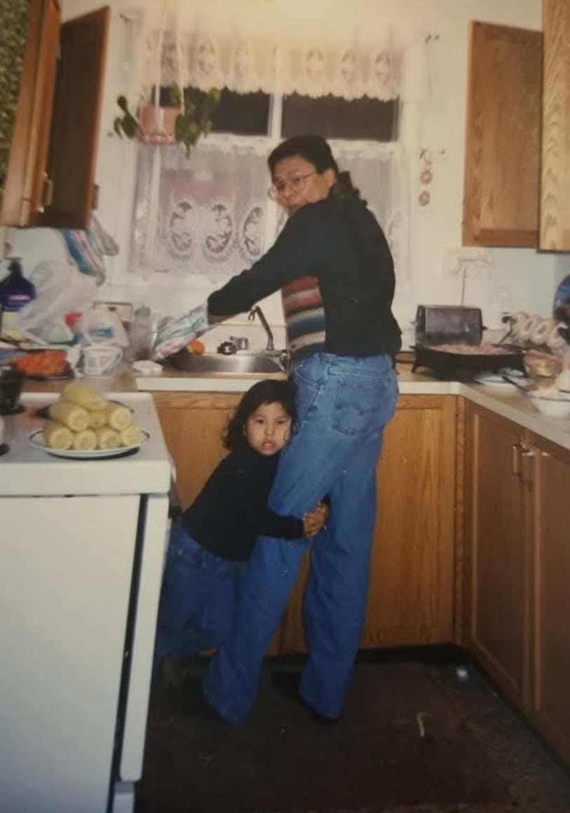 A boy clings to the leg of a man doing dishes in a kitchen.