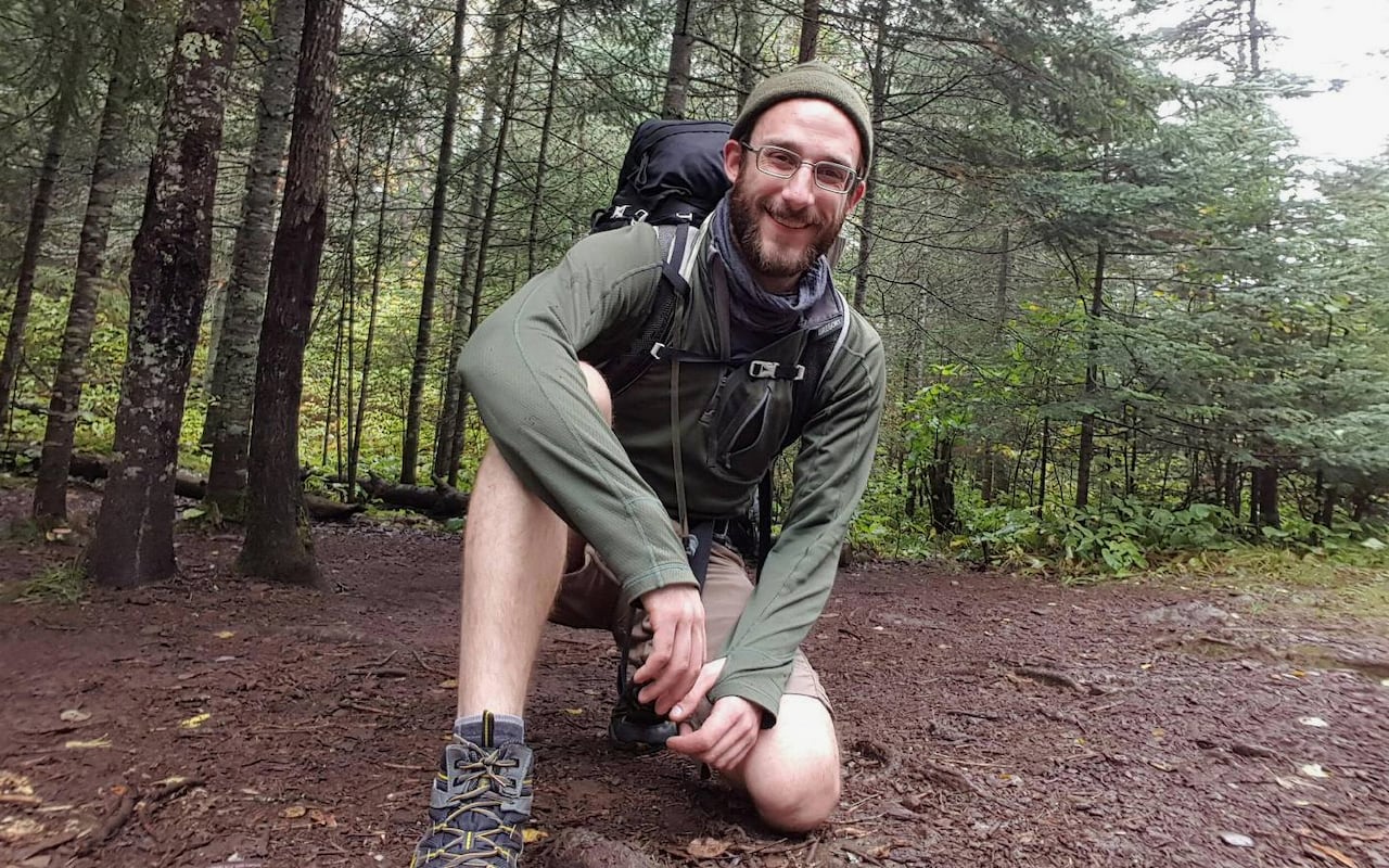 A man with glasses, wearing a green cap and shirt and a backpack, is shown in a forested area.