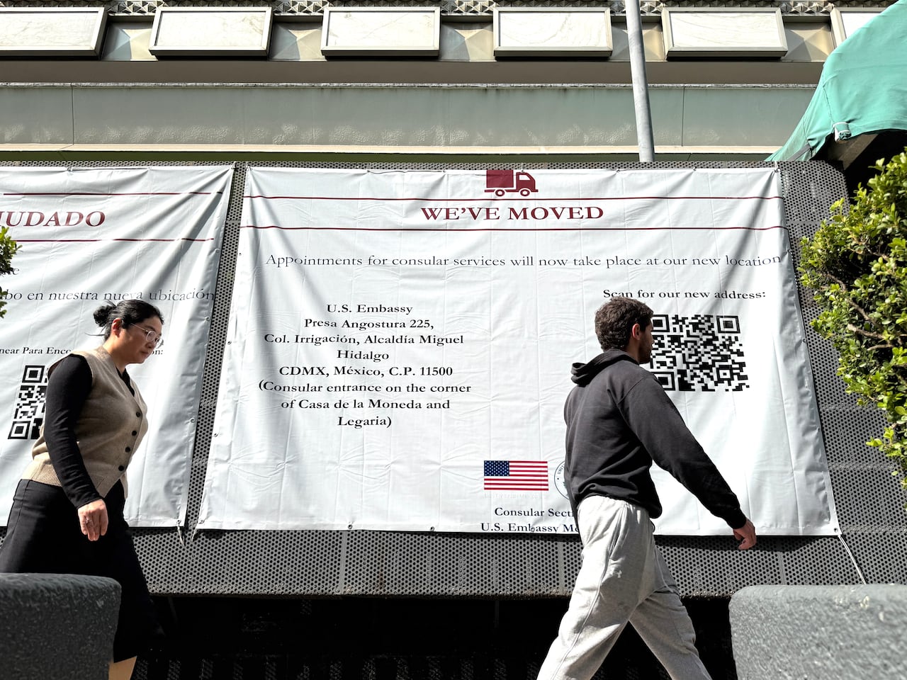 People walk past a large sign on the side of a building informing people that the U.S. Embassy in Mexico City has moved.