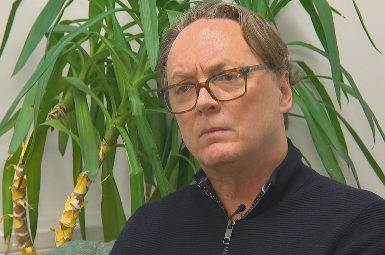 A man with glasses sits in front of a plant.