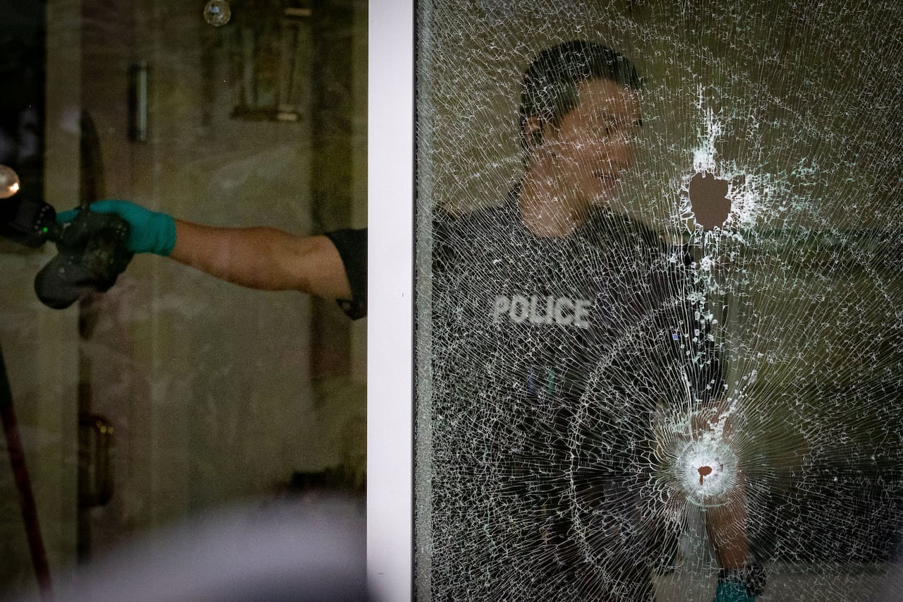 A police officer holding a camera in front of a window shattered by two bullets.