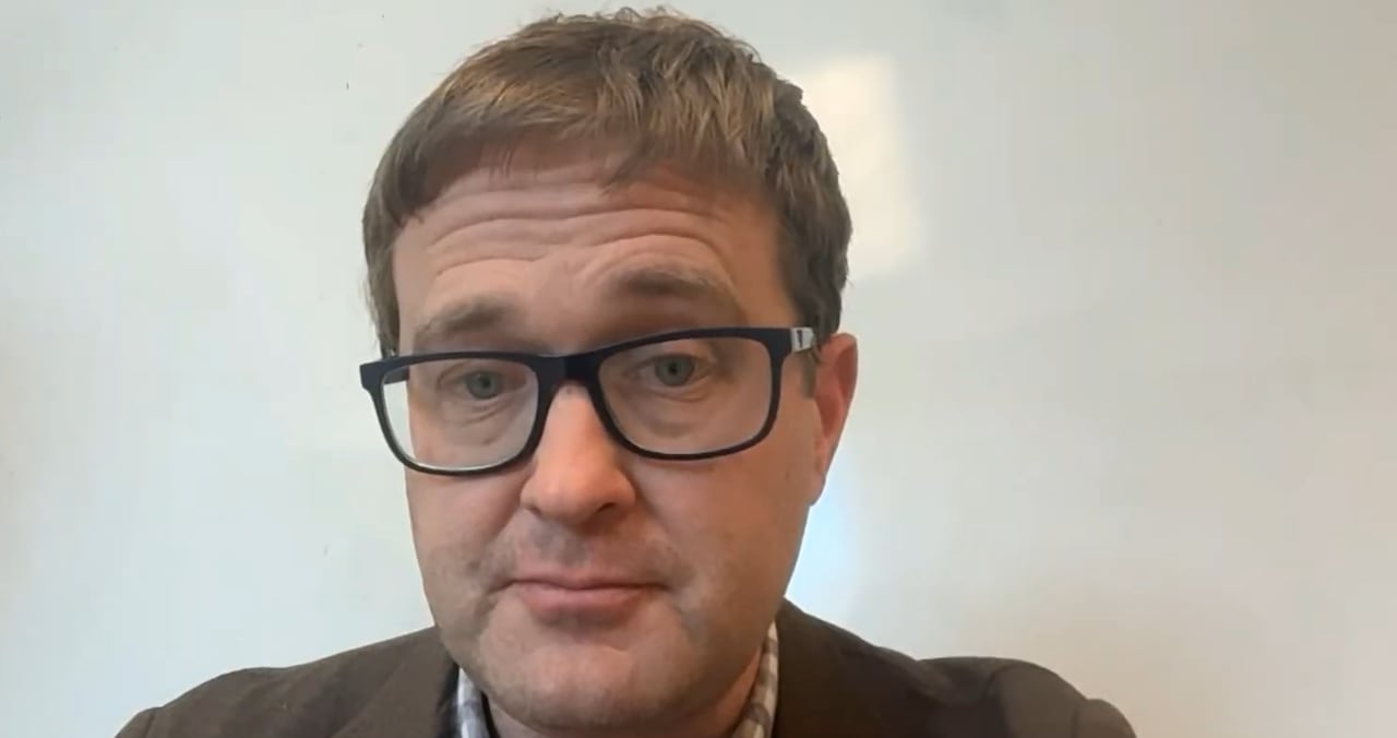 A man wearing glasses and a brown suit jacket sits in front of a white background.