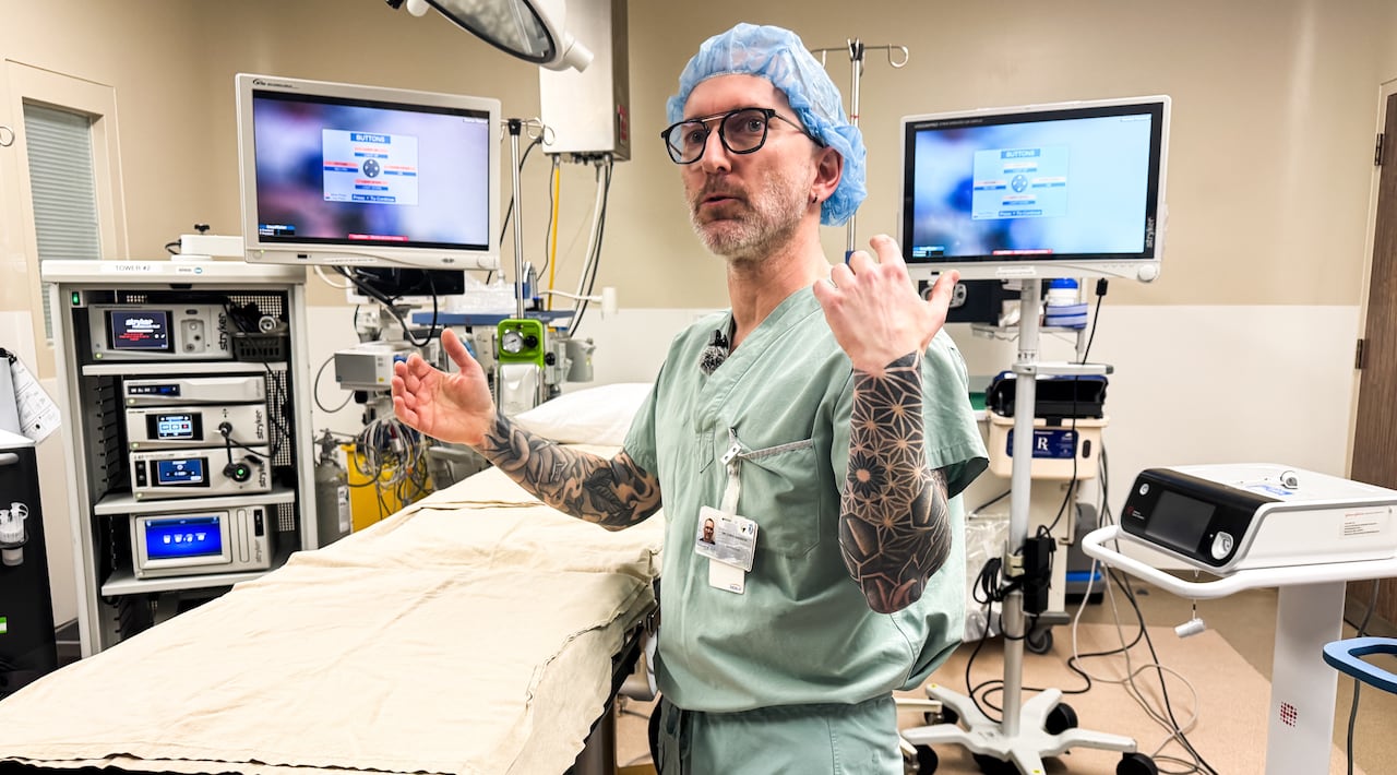 Man wearing blue surgery scrubs and a hair net with black tattoos on his arms is seen standing in a surgical suite with monitors and equipment
