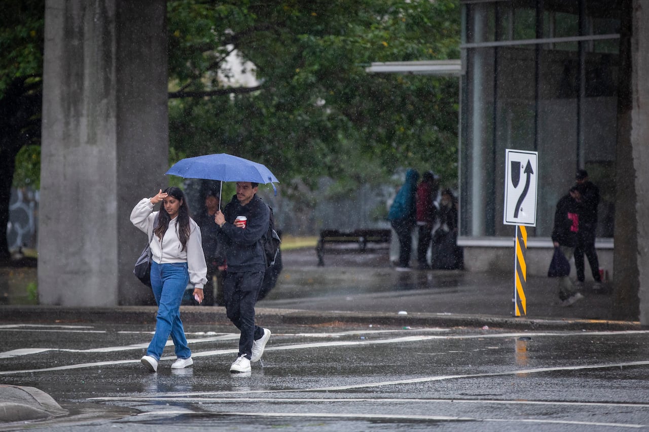 Two people cross a road while carrying an umbrella, while others mill around behind them.