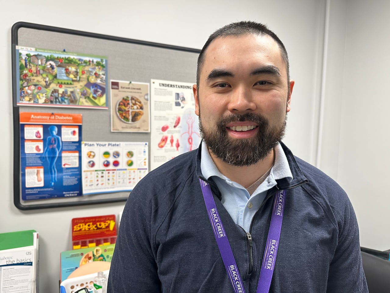 A man with a beard wearing a blue cardigan, shirt and purple lanyard. He is smiling in front of a bulletin board with food posters. 