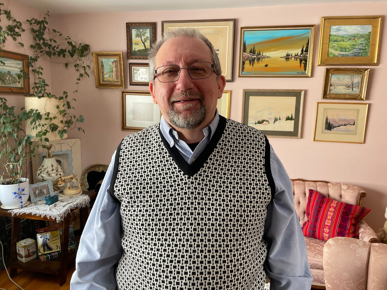 An older man in a sweater vest stands in a living room with photos on the wall and plant vines in the background.