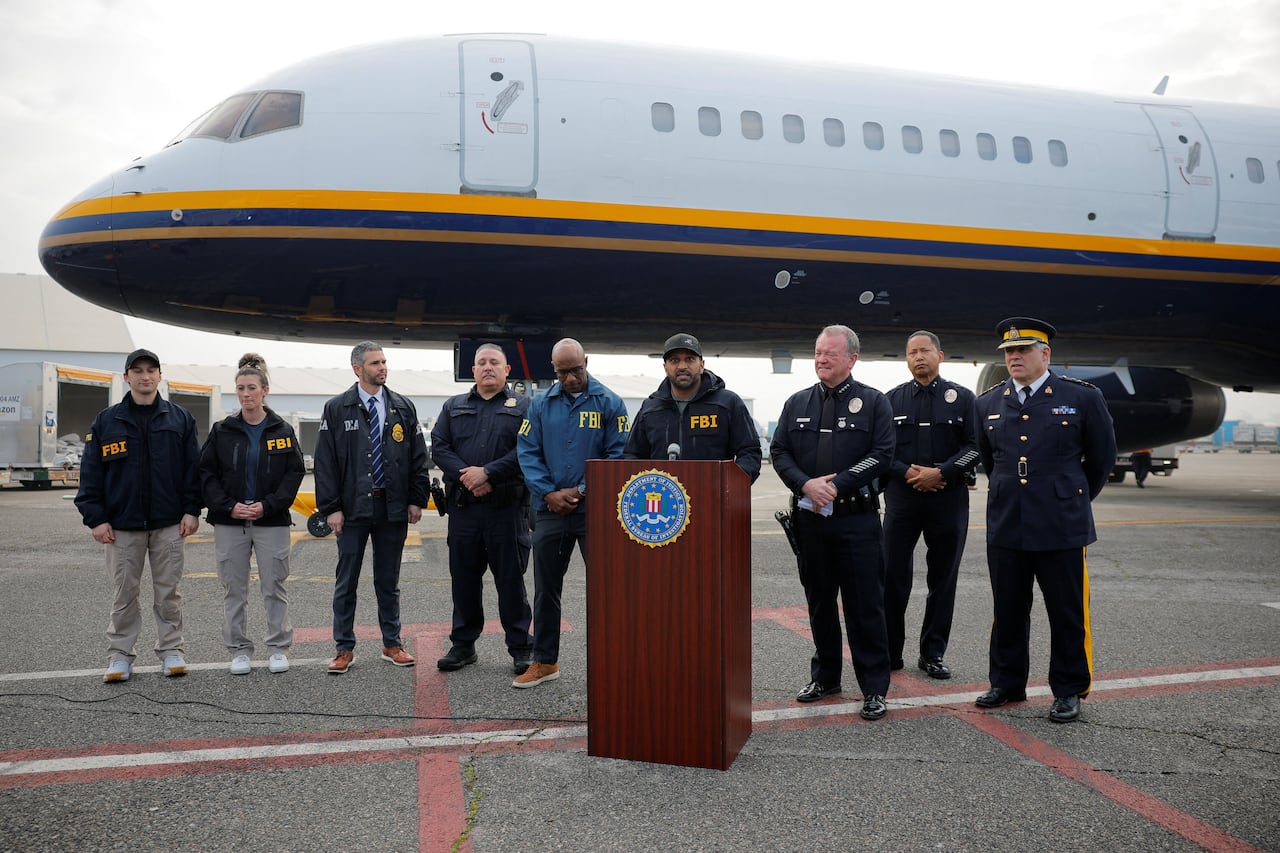 A man at a podium with other people behind him on a tarmac and a plane as backdrop during a news conference.