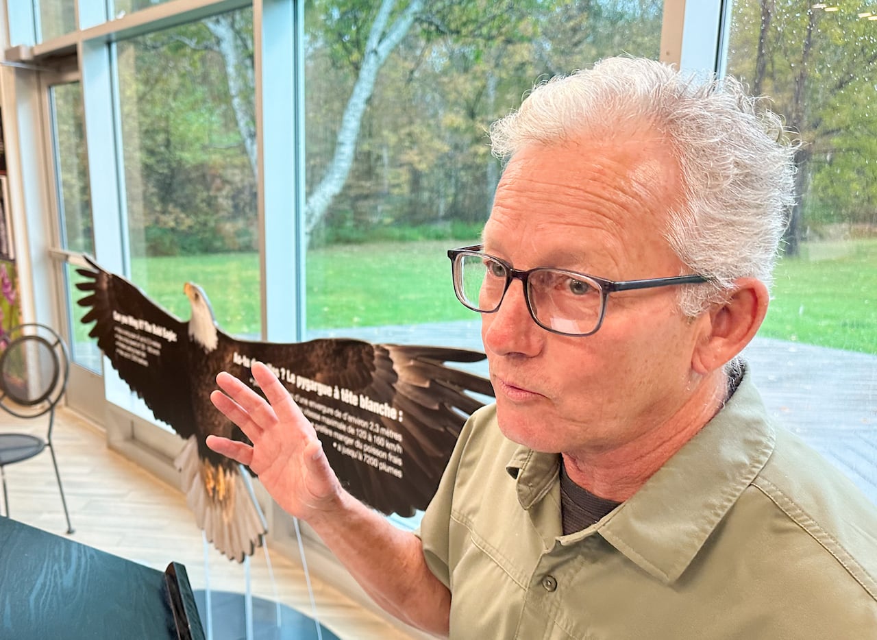 A man with white hair and glasses gestures with his hand while speaking in front of a window.