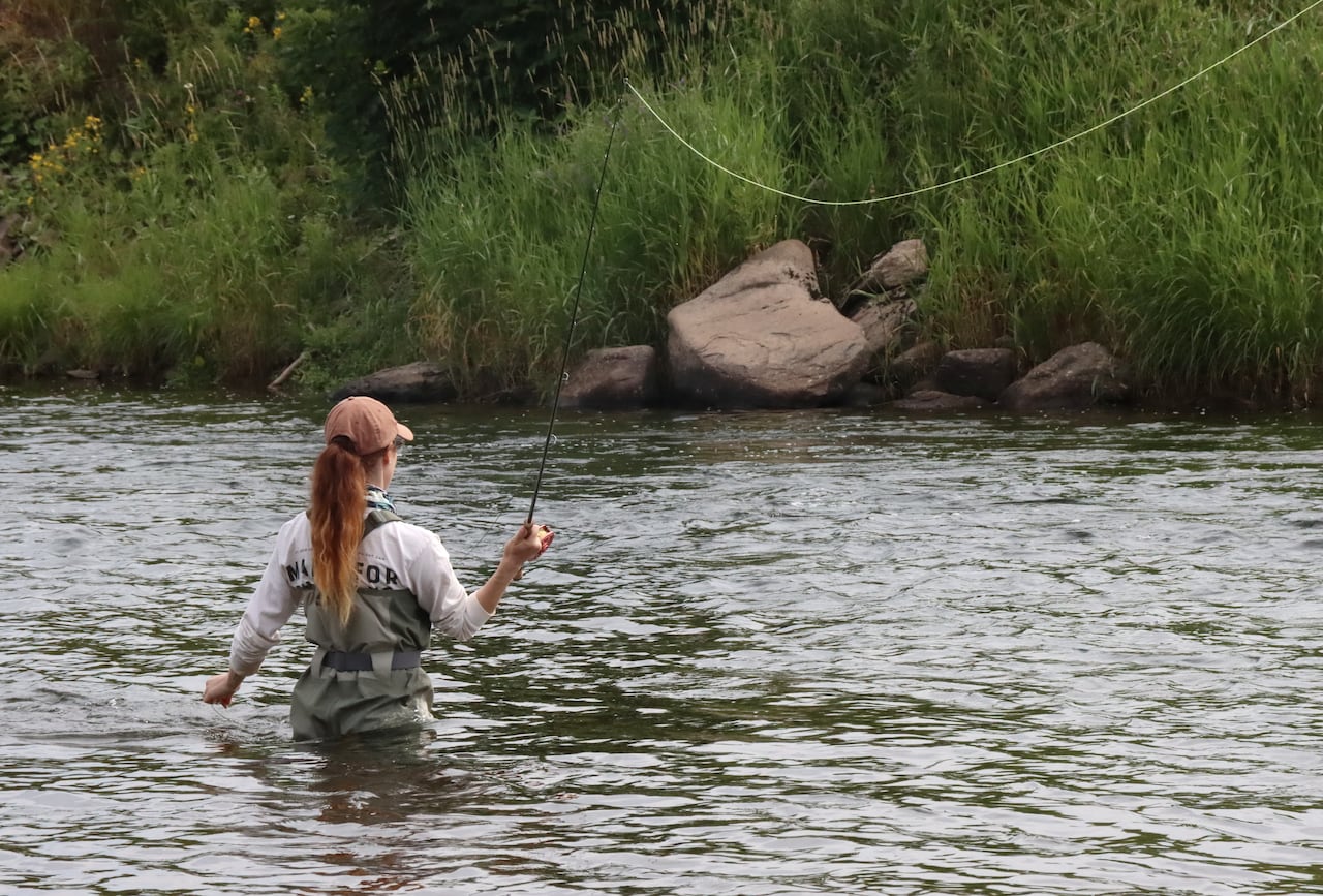 A woman stands in hip high river with her back to the camera. She casts a fly fishing line into the river in front of her.