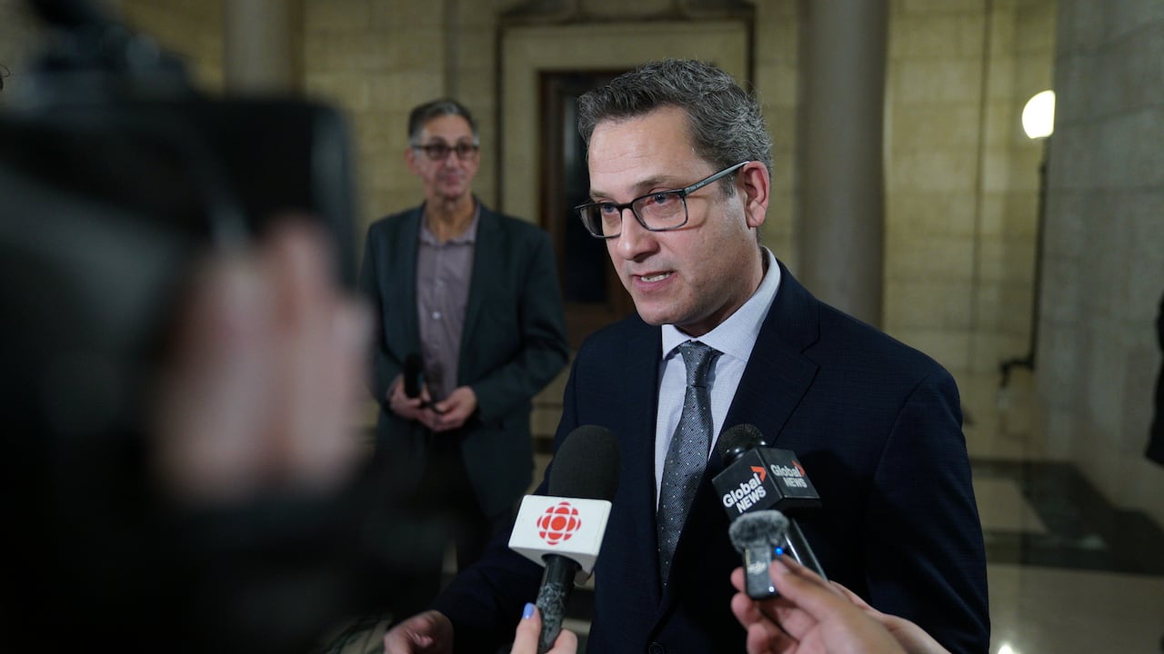 A man with brown hair and glasses in a suit speaks to reporters. 
