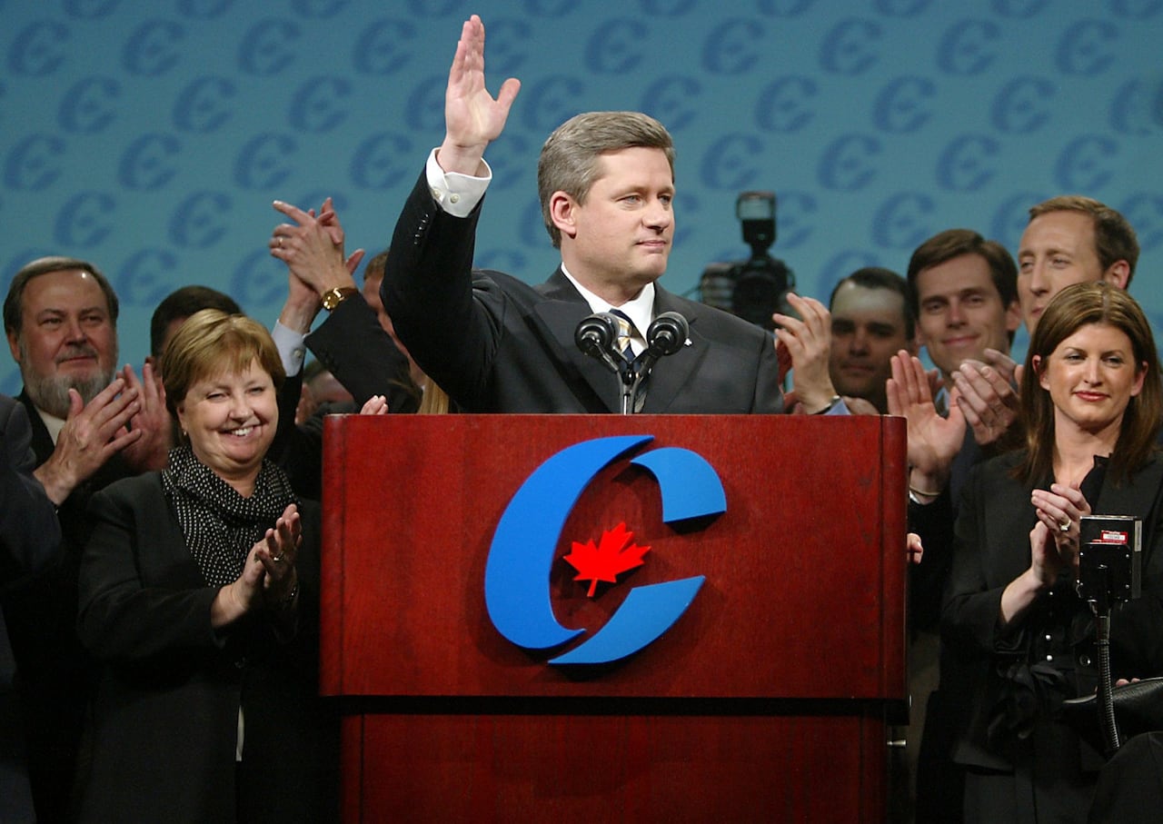 A man in a suit waves from a podium plastered with a large blue "C." People around him applaud.