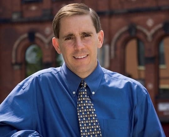 Man with brown hair in a blue shirt and tie stands in front of a brick building.