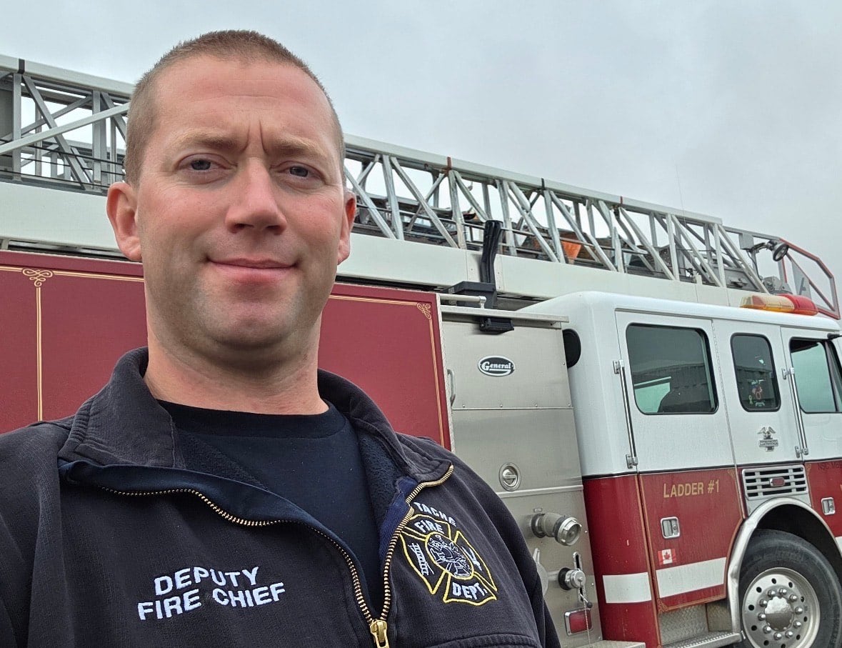 A man wearing a "deputy fire chief" jacket stands in front of a fire truck.