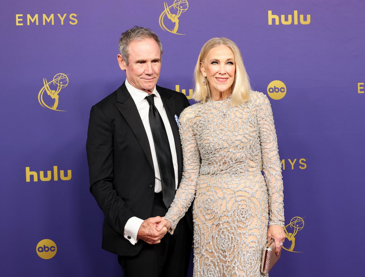 Actress Catherine O'Hara dressed in a silver gown holds hands with her husband, dressed in a suit, while they pose on a red carpet.