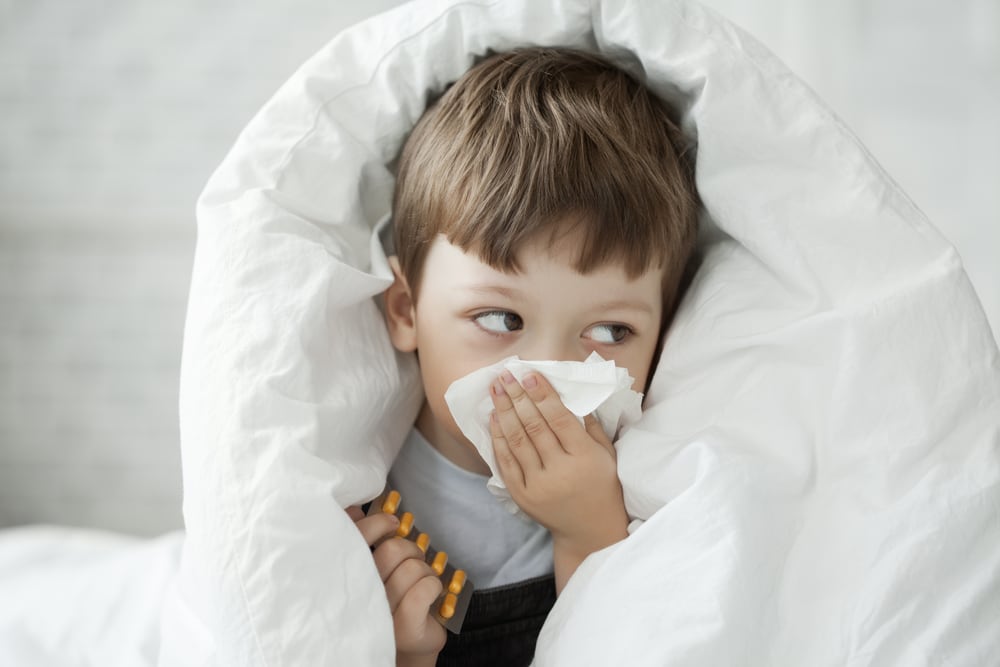 A boy wipes his nose with a tissue and is surrounded by a blanket.