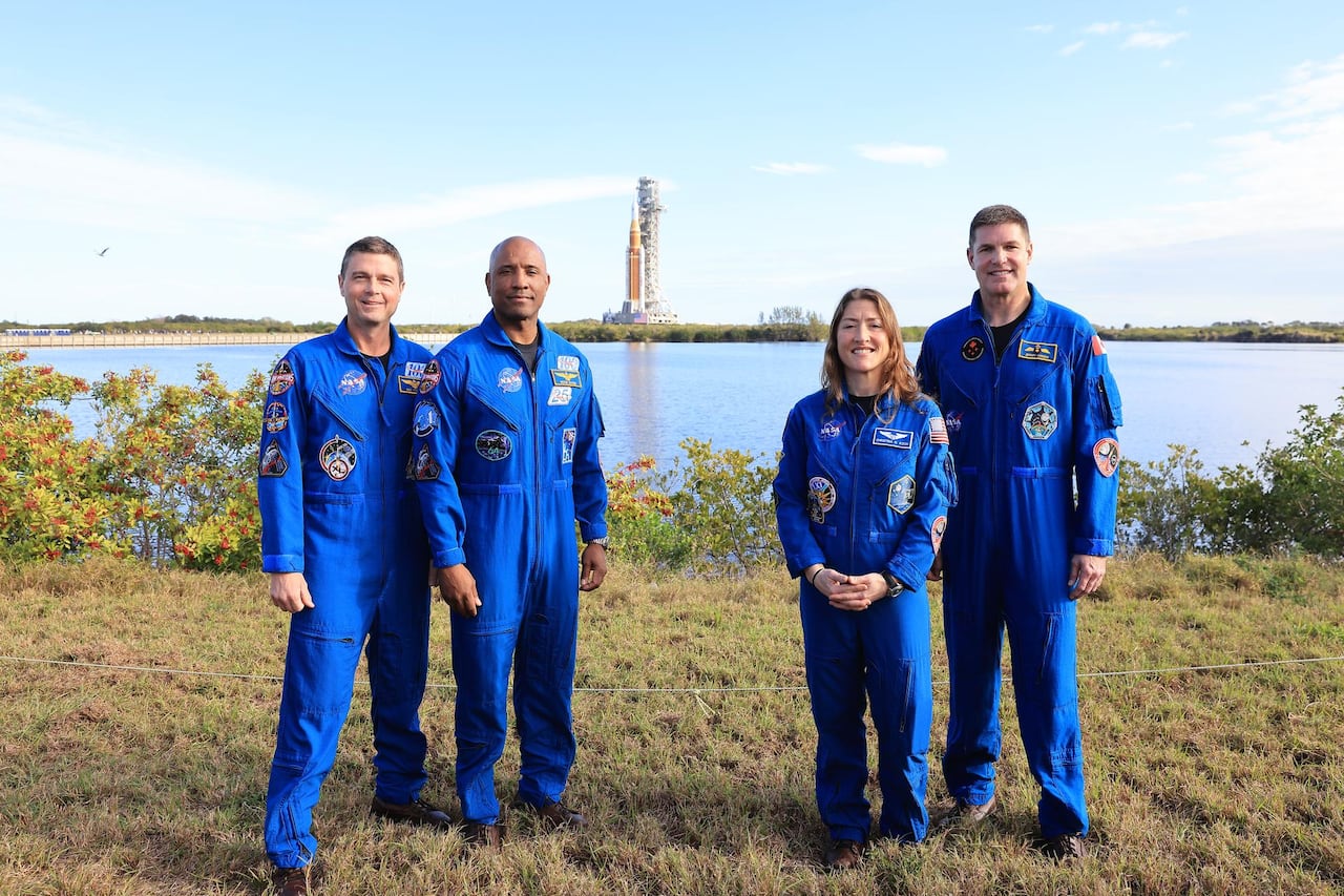 Four astronauts in blue spacesuits stand in the grass with an orange rocket in the background.