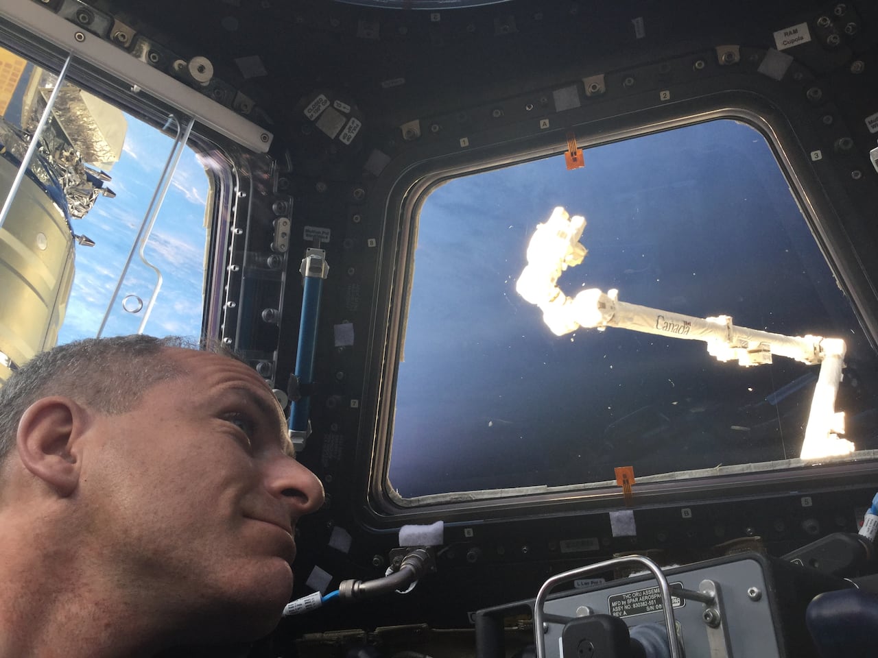 A close-up. of a man's face looking out the window into space with a robotic arm in the distance.