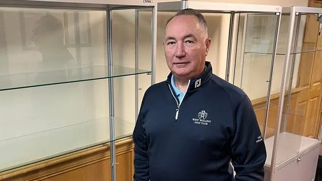 Vice captain of the West Malling club Mark Avery stands next to an empty glass cabinet with bare shelves. He is wearing jeans and a navy top and has grey hair.