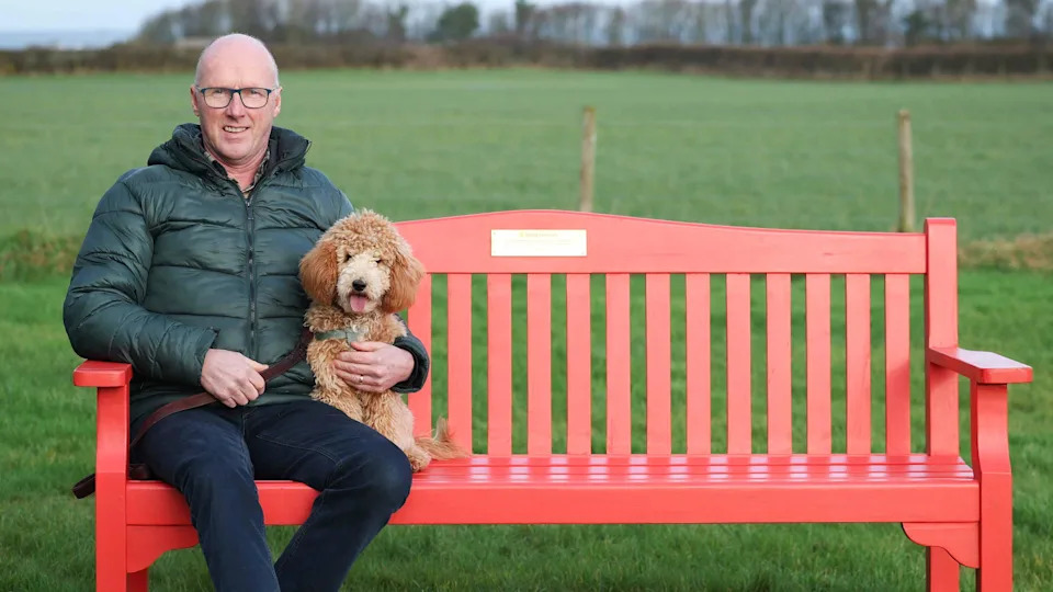 A man sitting on a red bench in the middle of a field. He is sitting beside a dog. He is bald and is wearing glasses, a dark green coat and dark denim jeans. 