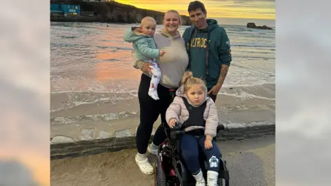 Charlie Brown Charlie Brown is stood shoulder to shoulder with his family, including Dani who is sitting in a wheelchair. They are posing for the camera, smiling. They're stood on a sandy beach and behind them is the coast, and shallow waves. Charlie has a green hoody on and short brown hair, next to him is a woman with a cream hoody and black leggings with white crocs on. She holds a baby with a mint green jacket and a white onesie with coloured dinosaurs on. 