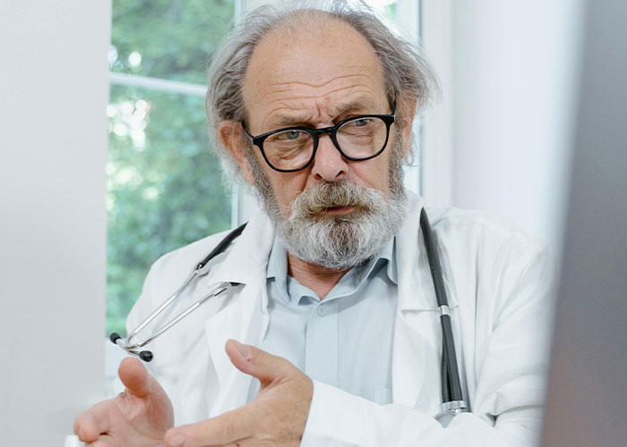 Elderly male doctor with glasses and stethoscope gesturing while discussing unprofessional things doctors have said.