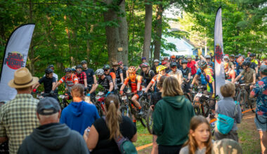 A crowd of mountain bikers is gathered for a race in a wooded area. They are lined up on their bicycles, wearing helmets and cycling gear, with a few spectators in the foreground. A banner with the event
