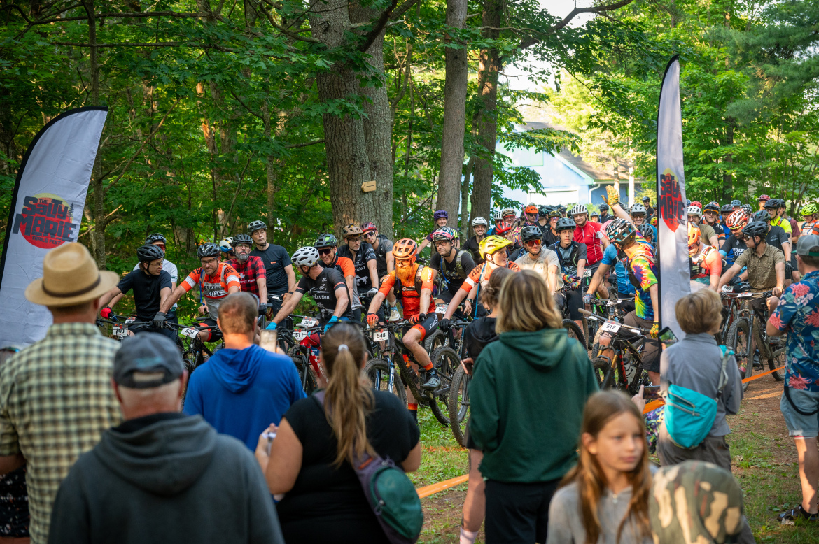 A crowd of mountain bikers is gathered for a race in a wooded area. They are lined up on their bicycles, wearing helmets and cycling gear, with a few spectators in the foreground. A banner with the event