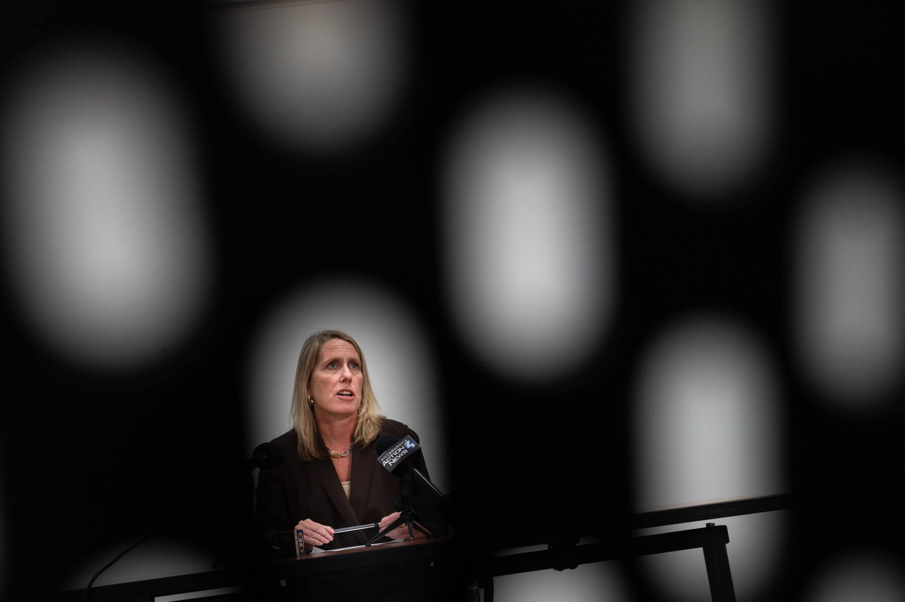 A woman stands at a podium speaking into a microphone, seen through an abstract pattern of blurred oval shapes in the foreground.