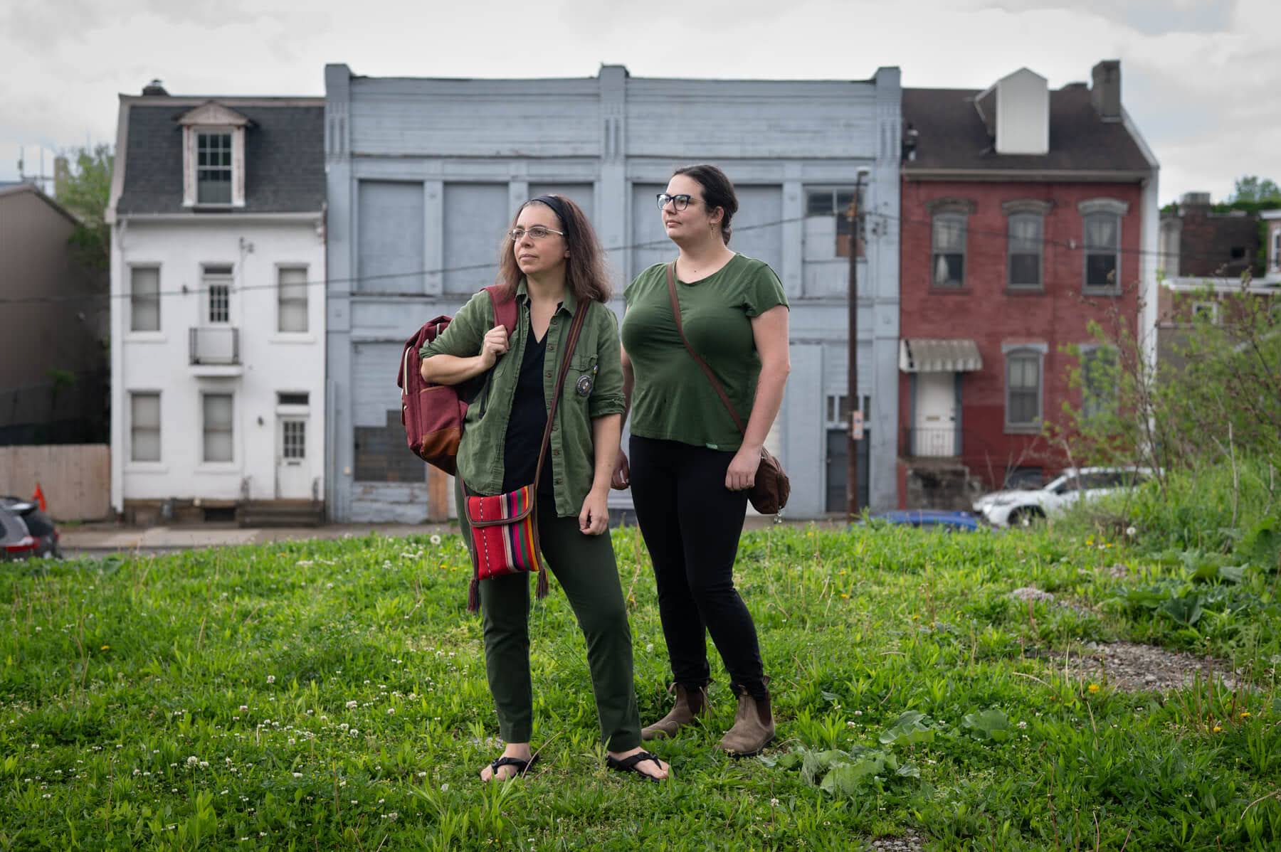 Two women stand on a grassy lot, looking off to the side, with older residential buildings in the background under a cloudy sky.