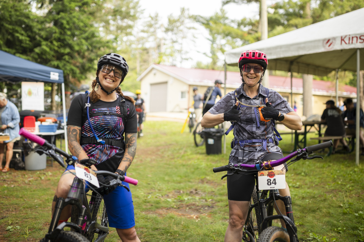 Two women smiling and posing on their mountain bikes in an outdoor setting, wearing helmets and athletic gear. One woman has a tattooed arm and is sitting on her bike with pink handlebars, while the other is giving a thumbs-up. In the background, there are tents and other mountain bikers preparing for an event.