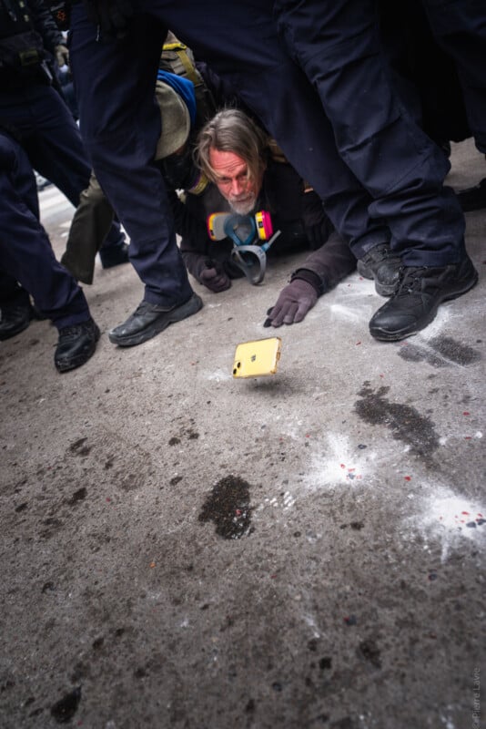 A man with a reddened face lies on the ground surrounded by people in dark clothing and boots, while a yellow phone rests on the pavement in front of him.