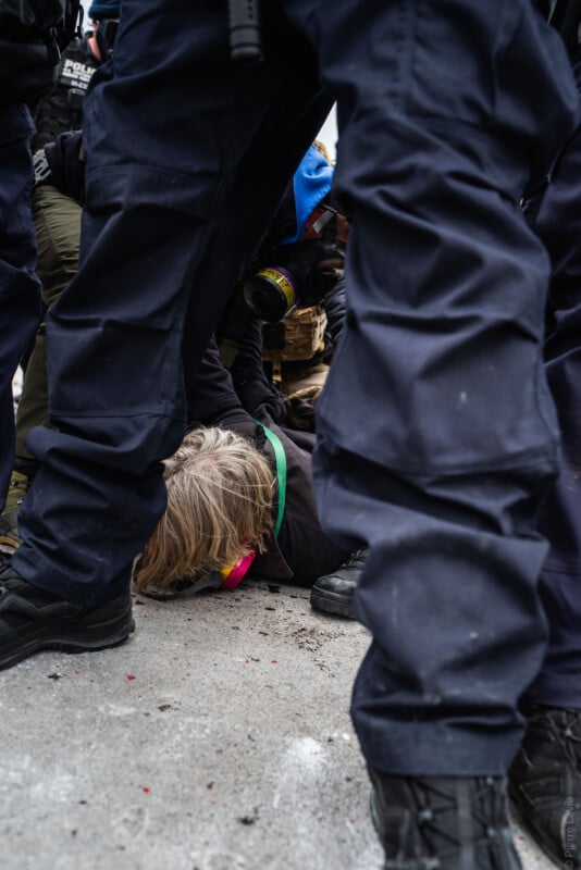A person with blonde hair lies face down on the ground, surrounded by police officers in dark uniforms. The person wears a green lanyard and pink ear protection, and is partially obscured by the officers’ legs.
