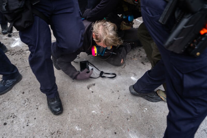 A person is pinned to the ground by several police officers on a concrete surface. The individual is holding a camera and appears restrained, surrounded by officers in dark uniforms.