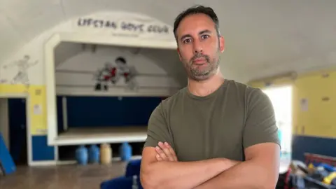 Stuart Woodward/BBC Jay Solder stands inside the Lifstan Boys Club building. He wears a dark green T-shirt, and he has dark short hair and a stubbly beard. He stands with his arms folded across his chest. Behind him is a stage with a boxing ring in the middle, and above the stage are the words "Lifstan Boys Club".