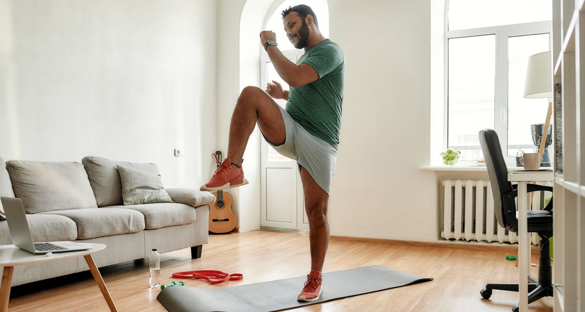 man wearing tshirt and shorts exercising in a living room setting standing on an exercise mat performing high knee to opposite elbow move.