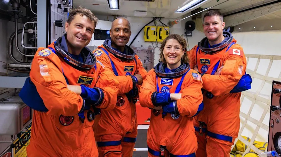 Four astronauts wearing bright orange spacesuits but no helmets. From left to right they are Reid Wiseman, Victor Glover, Christina Koch and Jeremy Hansen. They are standing together positioned in a row with their arms crossed. The room they are in is filled with technical equipment and cabling.
