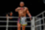 MMA fighter in colorful shorts in a ring, adjusts glove. Camera operator films nearby. Bright lights, focused expression.