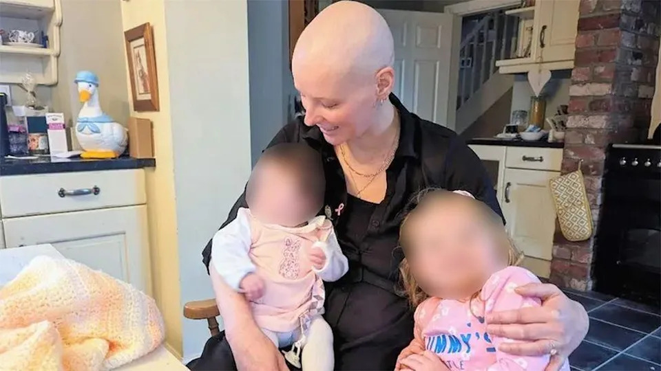 A mother, who has lost her hair as a result of chemotherapy, cradles an infant girl. She is sitting in an armchair a kitchen. Next to them is a young girl. She is  wearing a pink long-sleeved t-shirt. The faces of the children have been blurred.