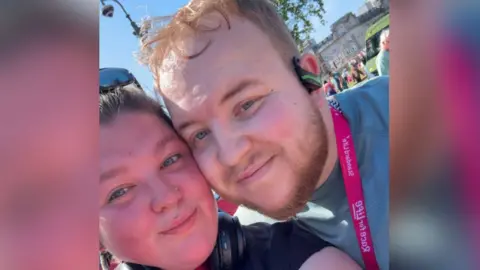 Richard Gallimore A close up selfie of Richard with his girlfriend, Sophie at the Race for Life running event. Sophie, on the left, has brown hair and wears sunglasses on the top of her head. Richard is standing next to Sophie with his cheek touching her cheek. He wears a green t-shirt, a pink medal, earphones and has short blonde hair. 