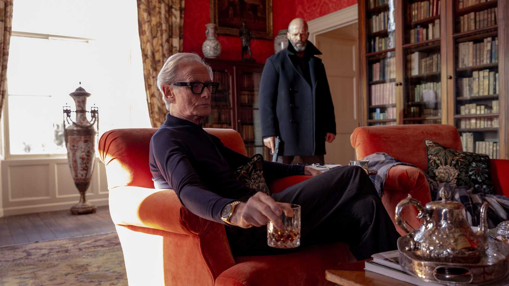 Bill Nighy, as Shelter's Big Bad, sits in a vividly orange plush chair, holding a cocktail, in a library-like room full of bookshelves and fancy vases, as Jason Statham looms behind him with a gun