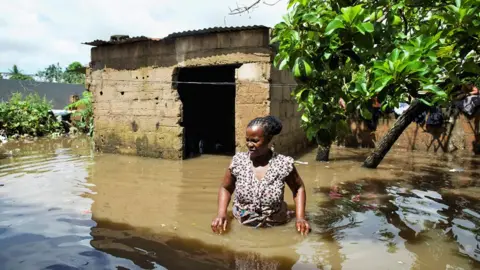 Reuters A woman standing with water up to her waist with a flooded building and tree behind her.