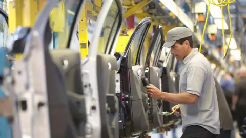 Getty Images A man in a grey tshirt and cap inspects a row of grey car doors inside a factory.