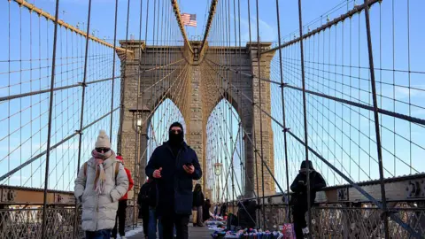 AFP via Getty Images Pedestrians walk across the Brooklyn Bridge in New York City. One woman wears a beige puffer coat, a wooly scarf, sunglasses and a wooly hat with her hands in her pocket. Next to her, a man is dressed in a black balaclava, long black puffer coat and trousers. Around them, the suspension cables of the bridge connect to a pylon with the United States flag flying atop it. The sky is clear and blue.